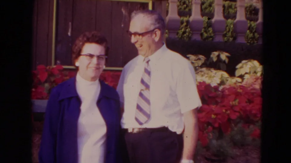 1974 Orlando home-movie frame of an older couple in glasses posing outdoors before red poinsettias and a dark building wall.