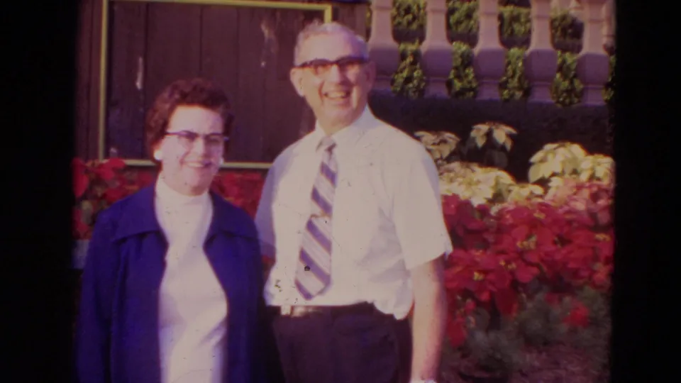 1974 Orlando frame showing a smiling older couple standing close together before poinsettias and a decorative exterior wall.