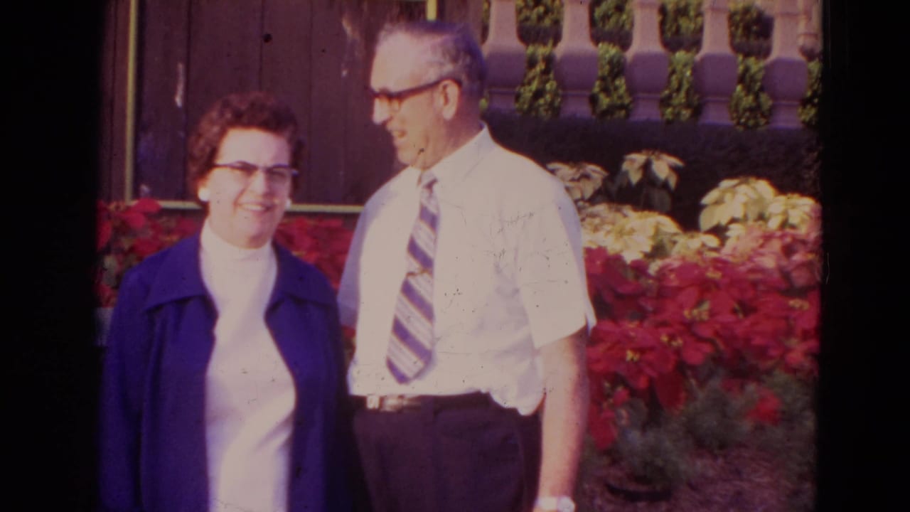 Elderly couple standing beside red flowers and a wooden fence.