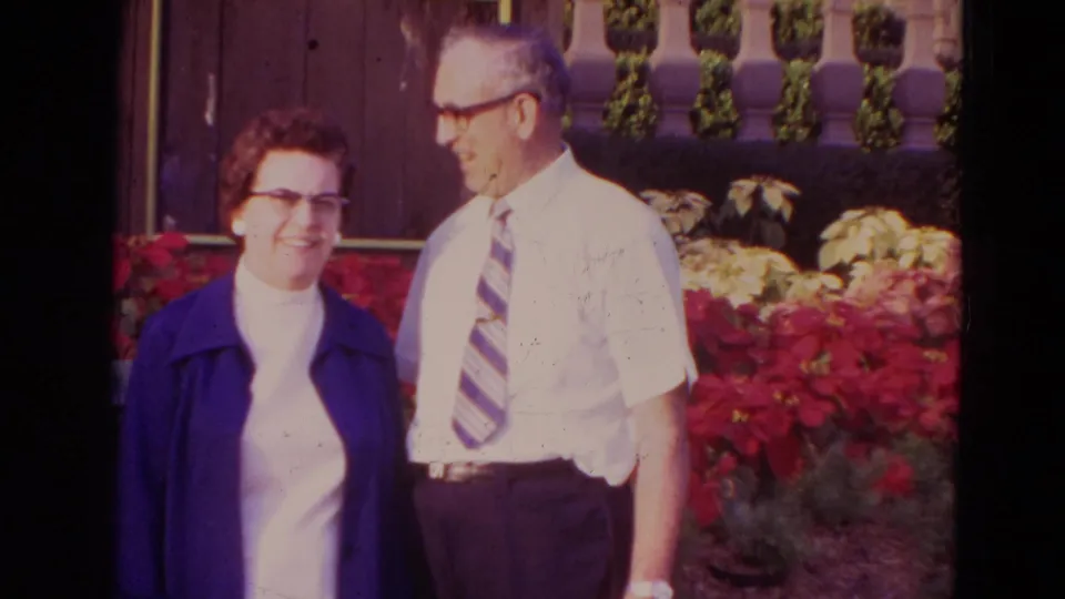 In supported 1974 Orlando home-movie footage, an elderly couple poses near red flowers, with the woman in a blue jacket and the man in a white shirt and tie.
