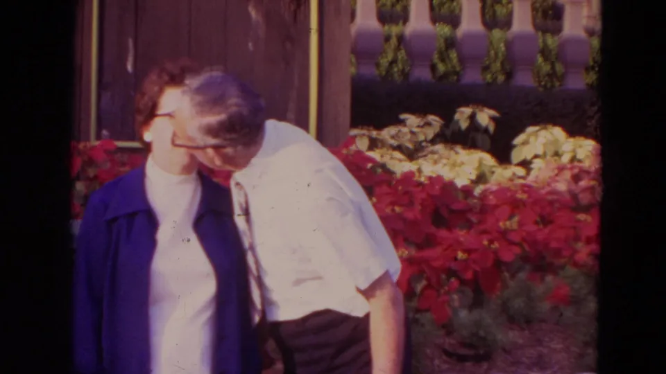 The elderly man leans in toward the woman while they stand in a sunlit Orlando garden with red flowers behind them.