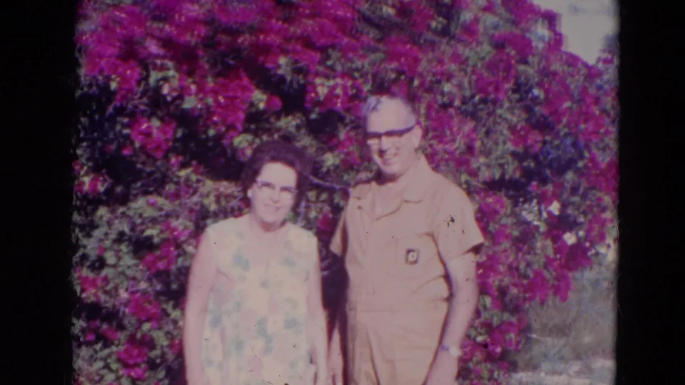 The couple stands together in a second Orlando garden setting, with purple bougainvillea behind them and the man wearing a light-colored jumpsuit.