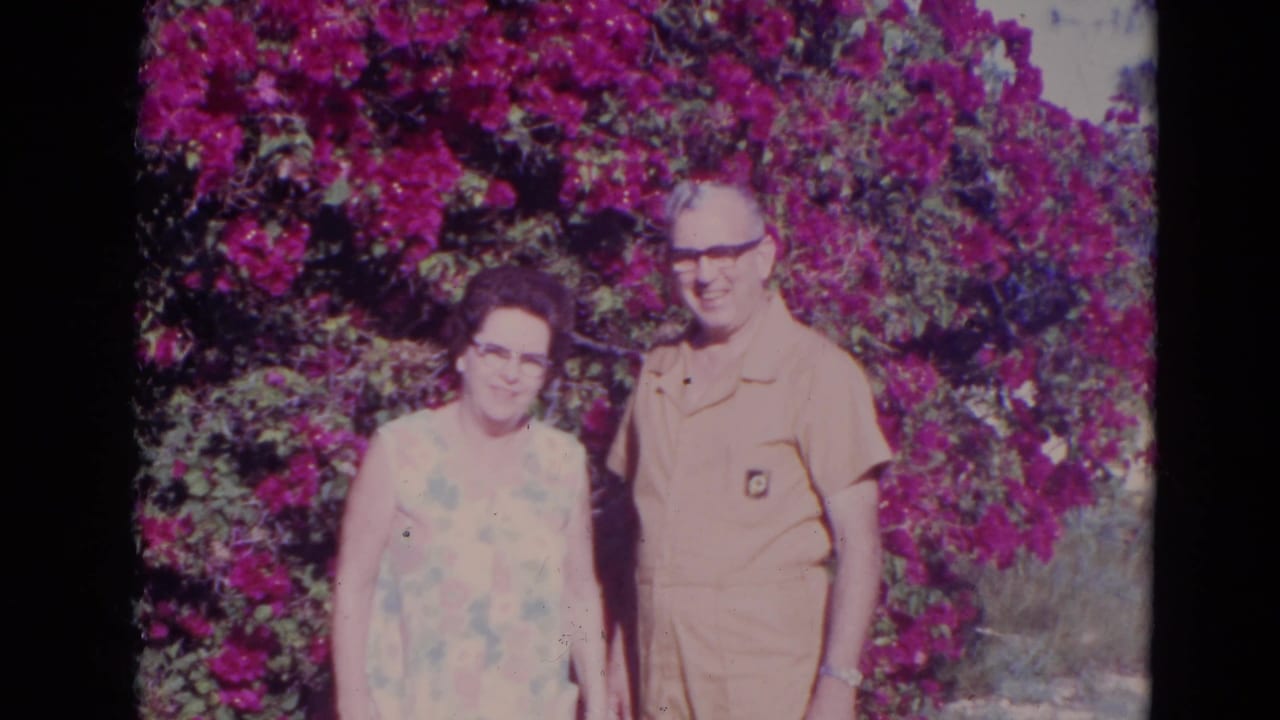In supplied 1974 Orlando 8mm footage, an elderly couple stands in sun before dense purple flowers and greenery.