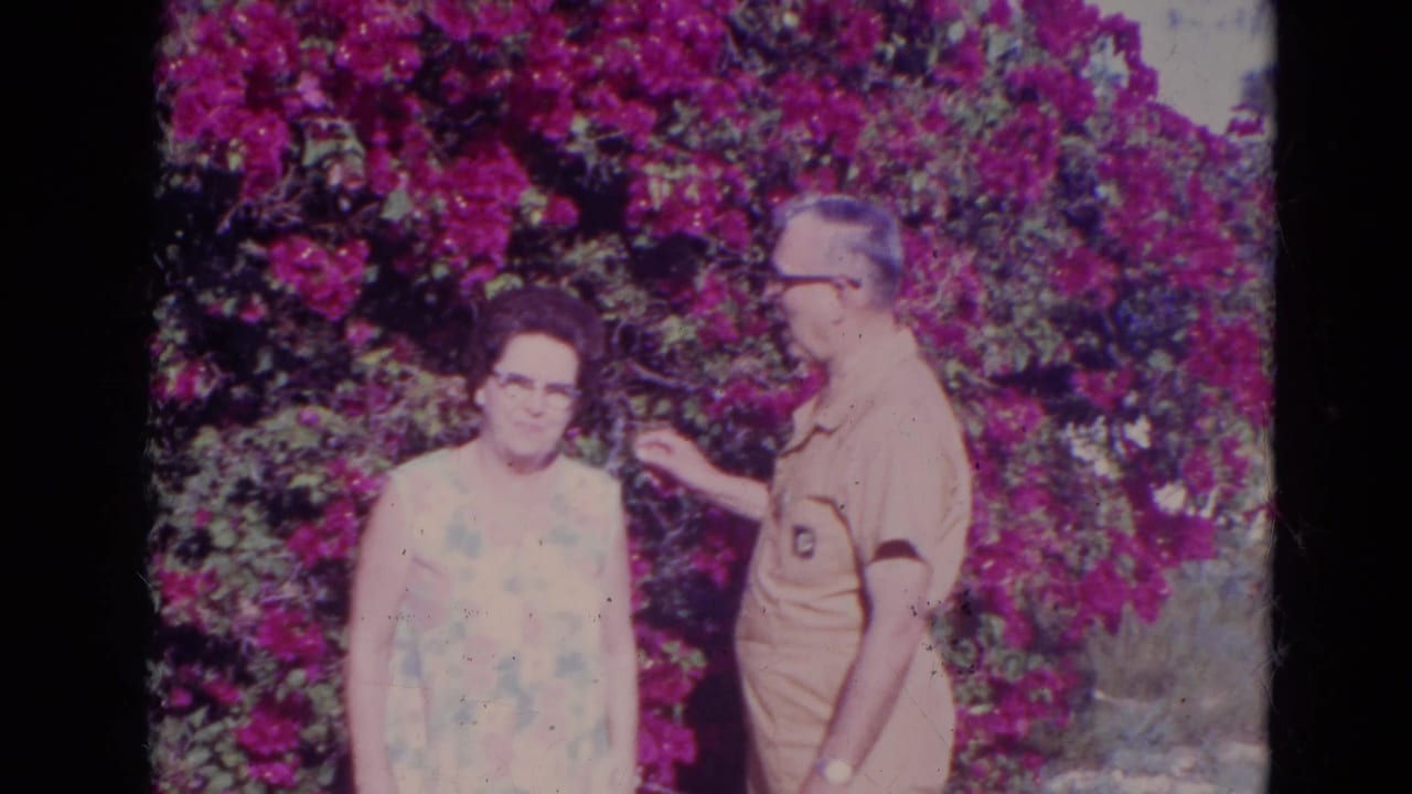 The woman looks toward the camera as the man in a tan outfit turns and reaches near purple flowers in the garden.