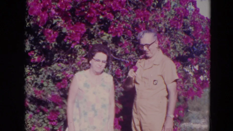 In 1974 Orlando home-movie footage, an older woman and older man stand beside vivid bougainvillea in a sunlit garden.
