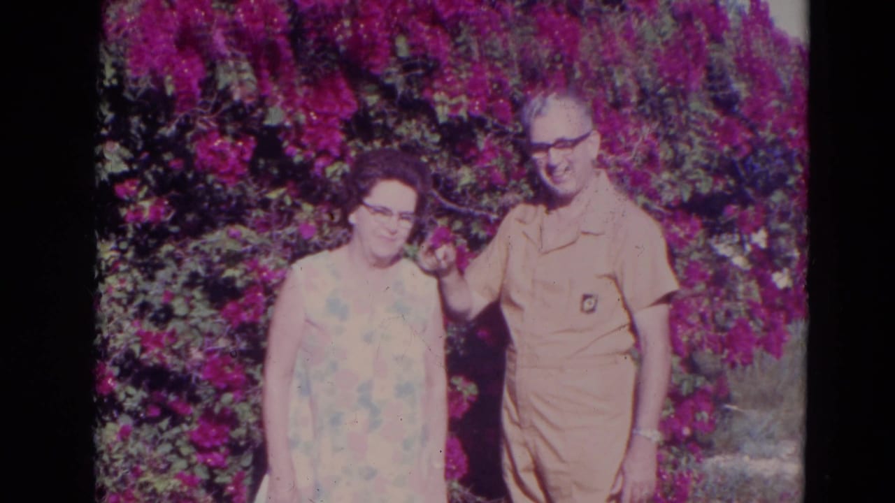 In the Orlando 1974 garden scene, the older man raises a small flower near the older woman as they stand close together.