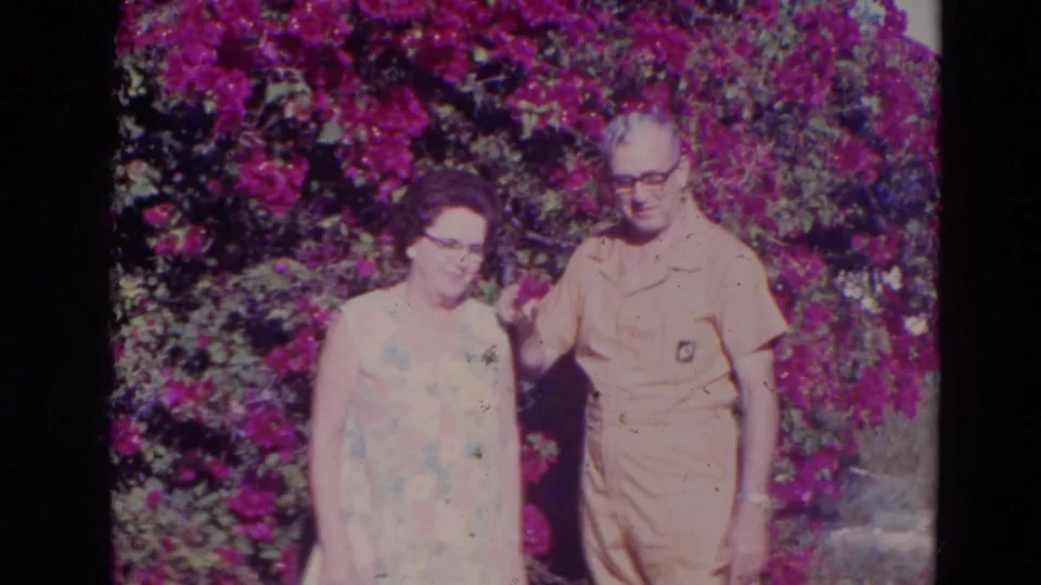 The 1974 Orlando home-movie frame shows the older couple close together, with the man’s hand near the woman and magenta flowers behind them.