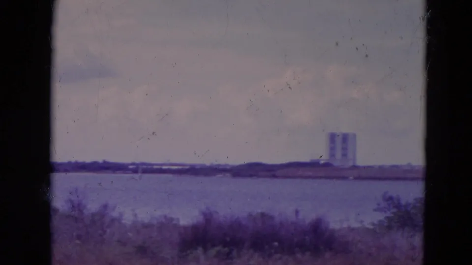 A brief 1974 Orlando-area waterfront view shows foreground grasses, water, a low shoreline and two distant tower-like buildings.