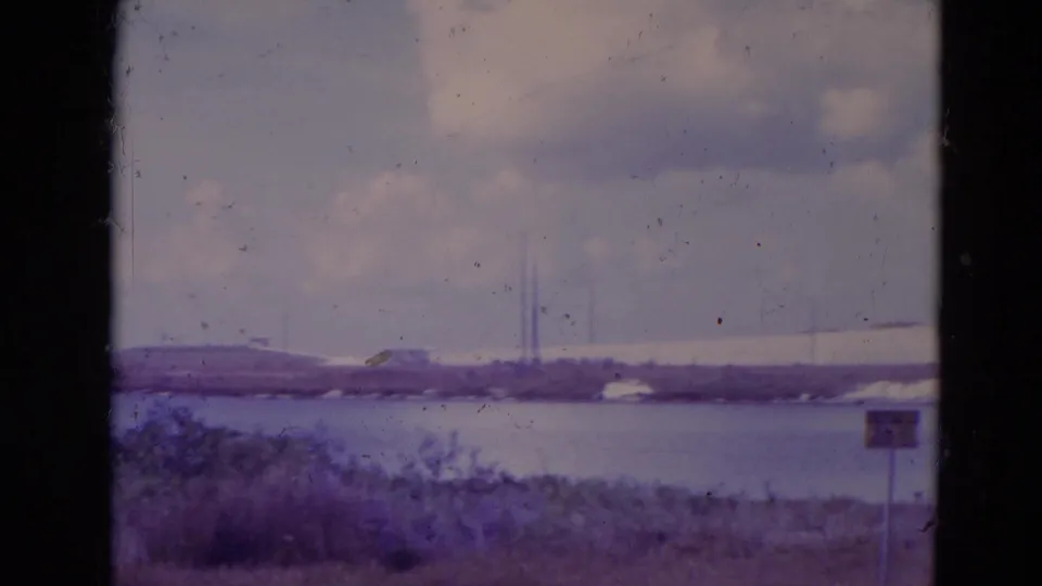 1974 Orlando-area 8mm frame showing water, distant low structures, foreground vegetation, a small sign, and cloudy sky.