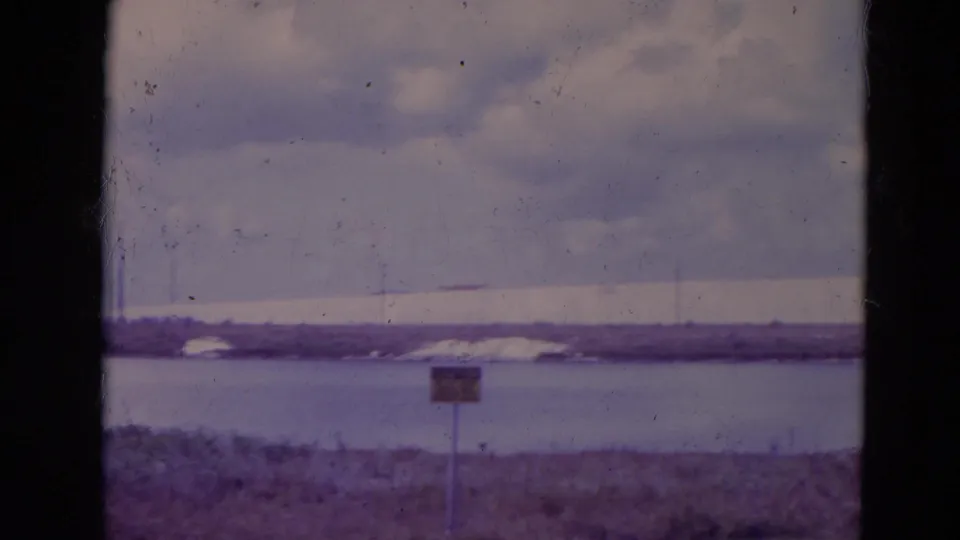 1974 Orlando-area 8mm frame showing a distant shoreline, low stadium-like structure, water, foreground sign, and film wear.