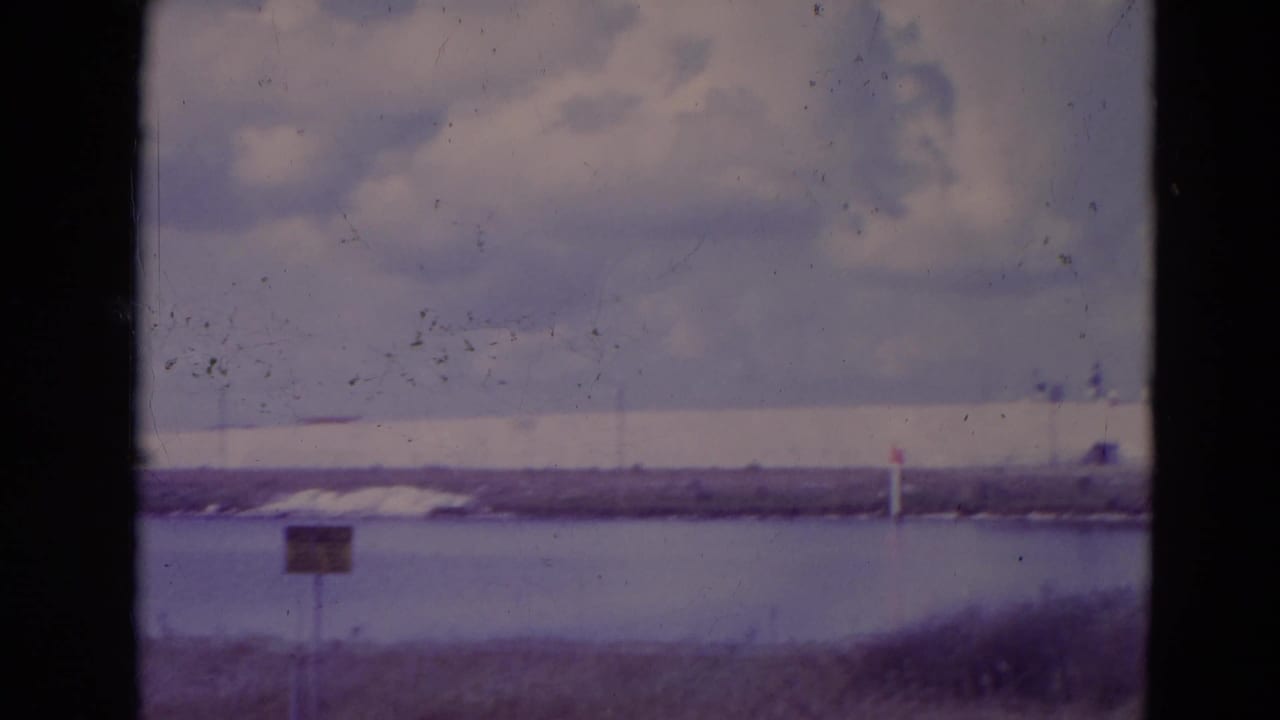 Waterfront view with clouds, grassy foreground, a small sign, and distant low structures across the water.