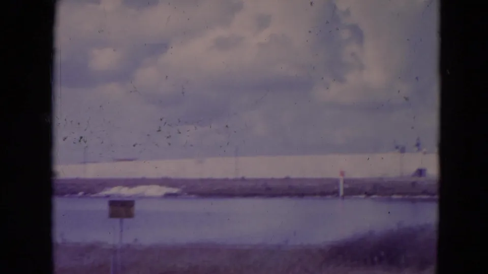 1974 Orlando-area 8mm frame showing water, grass, a small sign, clouds, and distant low infrastructure beyond the shoreline.