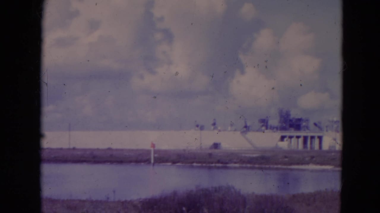 1974 Orlando-area 8mm frame with a distant white facility or low building across water under textured clouds.