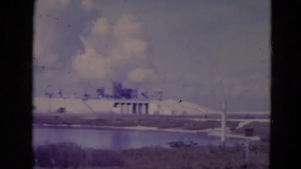 1974 Orlando-area 8mm frame showing a white waterside structure, grass, water, and distant low infrastructure.