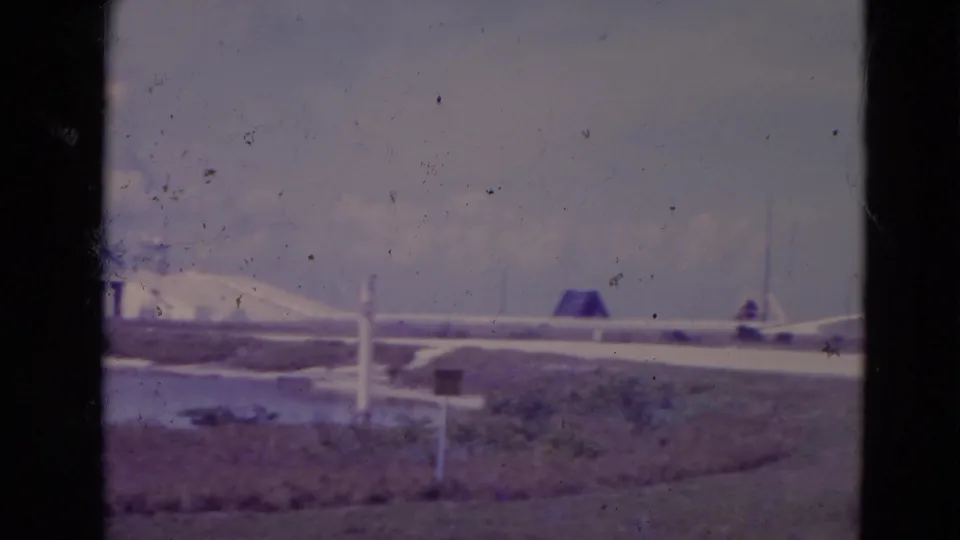 1974 Orlando-area 8mm frame with a runway-like area, grass, water, small signs, and a distant airplane-shaped form.
