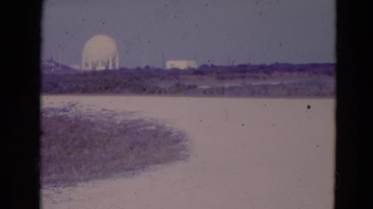 Distant white dome beyond flat scrubland and a pale channel under a hazy sky.