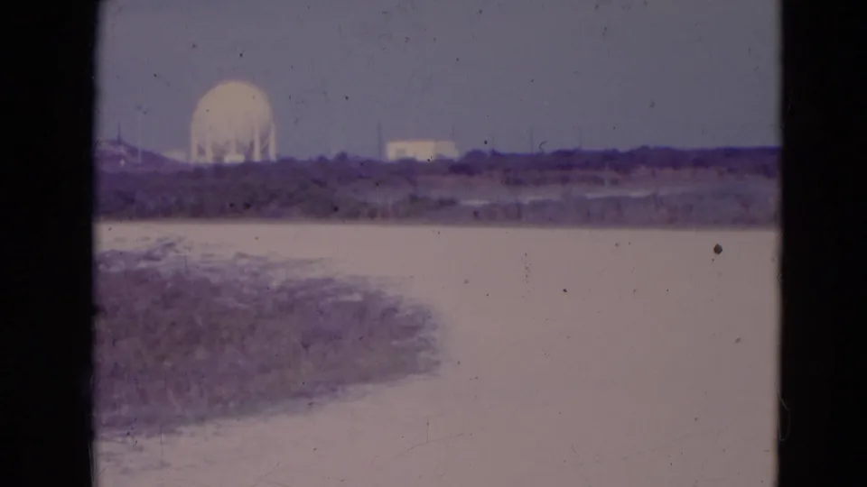 1974 Orlando-area 8mm frame showing a distant white dome, low structures, flat scrubland, and a pale water or road strip under a hazy sky.