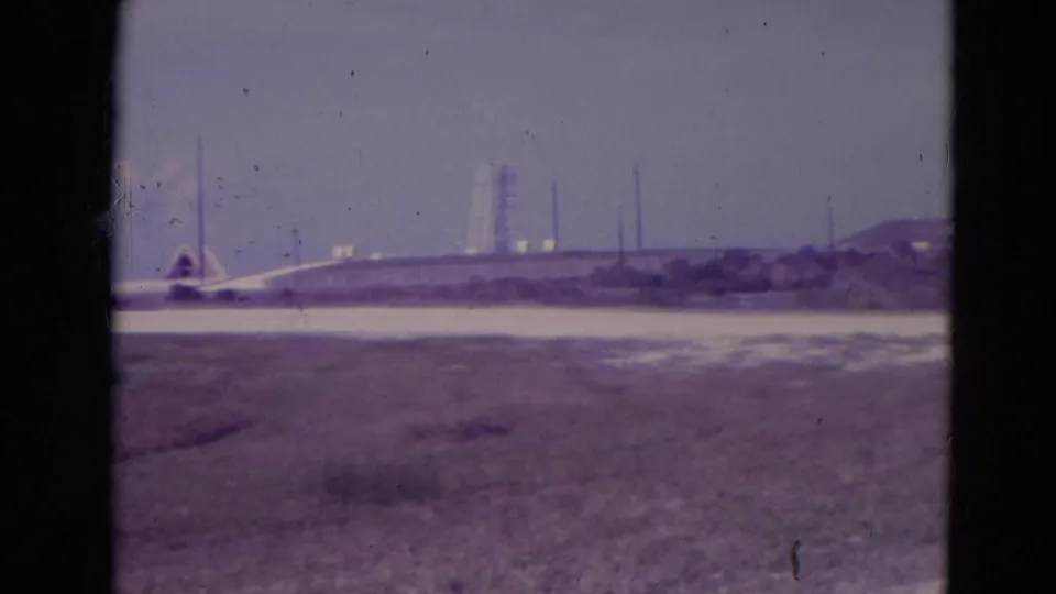 1974 Orlando-area 8mm frame showing distant industrial towers, low structures, open foreground terrain, and visible gate edges from the film scan.