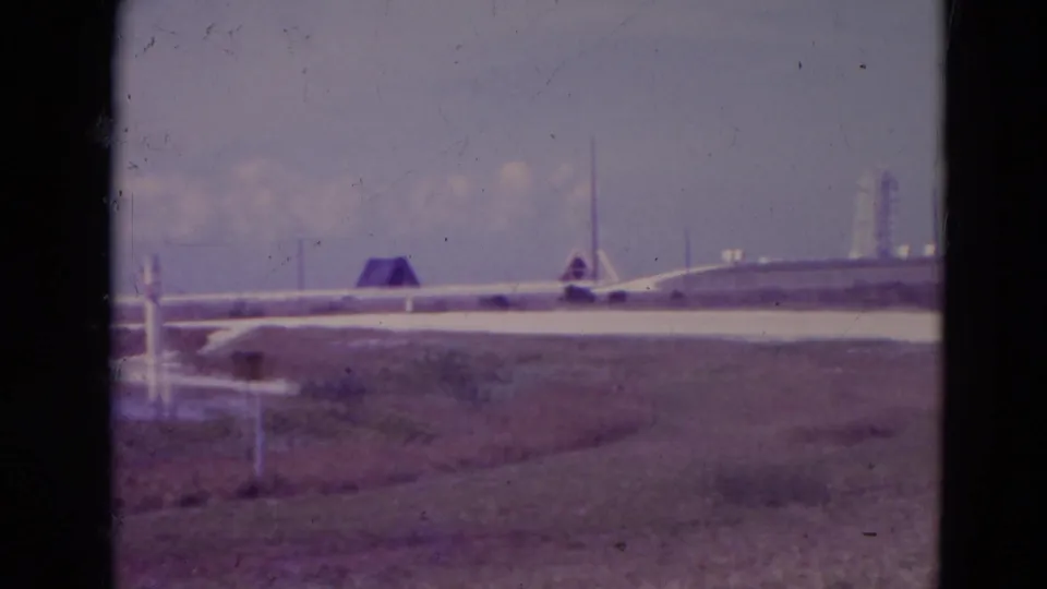 1974 Orlando-area 8mm frame showing a low industrial structure, a distant tower, pale channel or roadway, scrubby foreground, and hazy sky.
