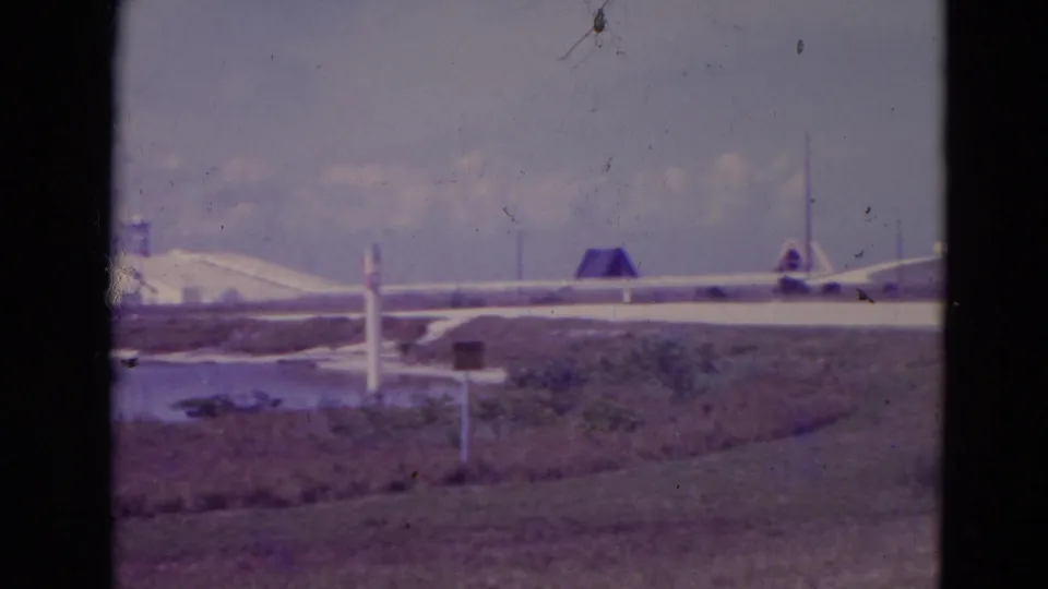 1974 8mm view toward Cape Canaveral launch pad structures across marshland and water, framed with dark film borders.