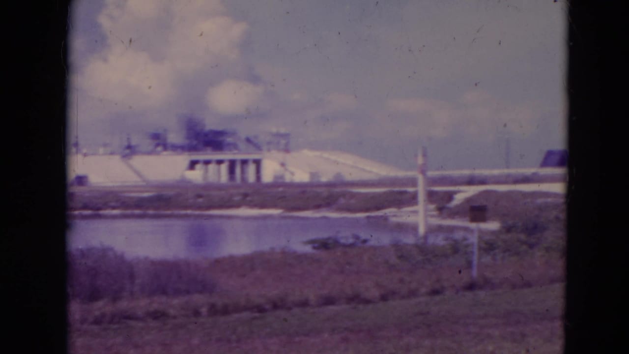 Visitor-shot 1974 view of Cape Canaveral launch pad structures beyond water and low marsh vegetation.