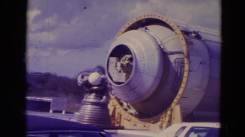 Close 1974 8mm view of a large rocket booster section at Kennedy Space Center, with cars and a small fixture nearby for scale.