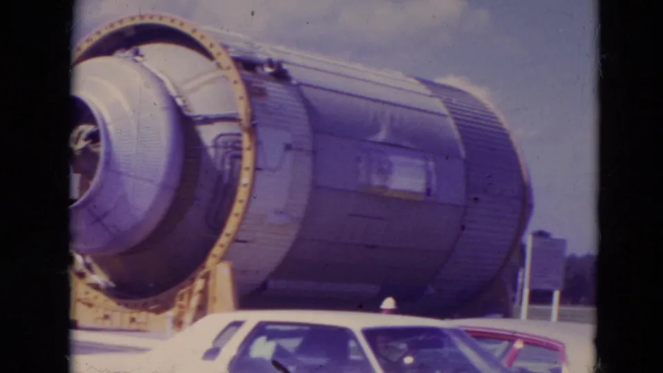1974 visitor-shot side view of a large rocket booster section at Kennedy Space Center, with parked cars in front.