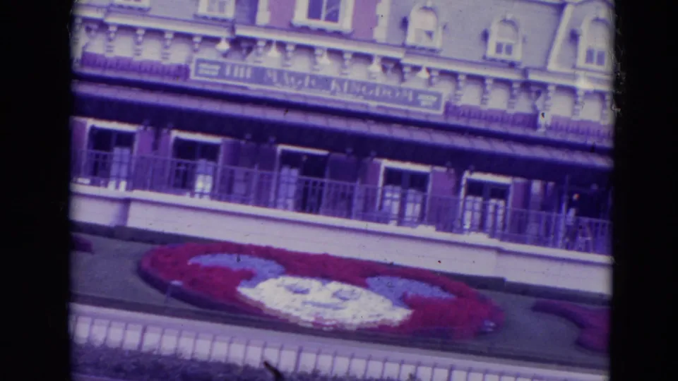 1974 Orlando home-movie frame showing the Magic Kingdom entrance building, balcony, sign band, and Mickey-shaped flower bed.