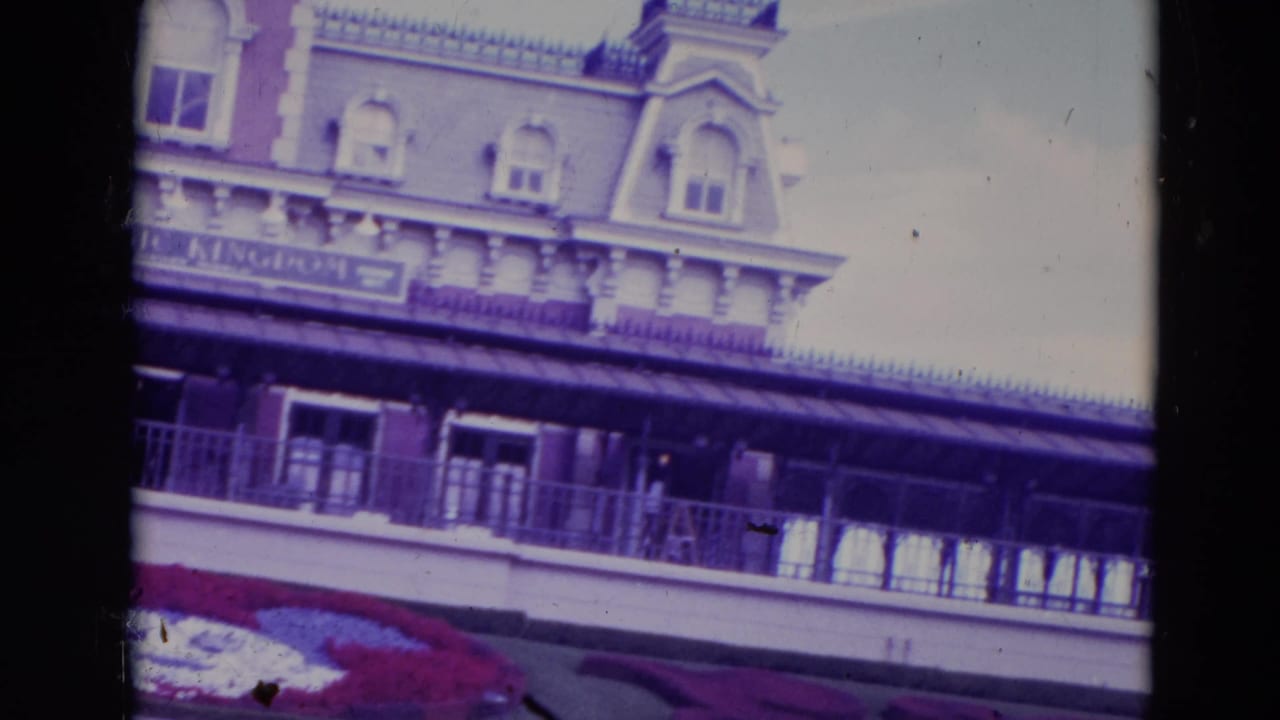 Entrance building with balcony, sign band, flower bed, and pale sky.
