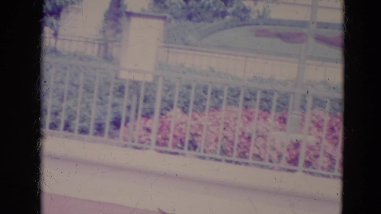 1974 Orlando home-movie frame showing a white fence, pale park architecture, and red floral shapes above the railing.