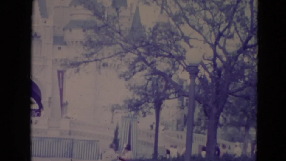 1974 Orlando frame showing Cinderella Castle beyond trees, a fence, lamppost, and small distant visitors near the walkway.