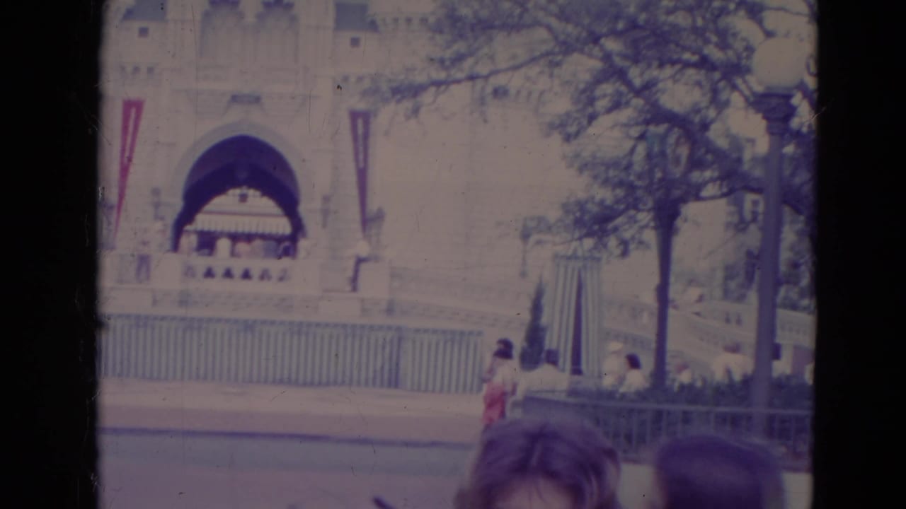 1974 Orlando home-movie frame showing a large pale arched facade, vertical banners, railings, trees, and partial visitors at the lower edge.