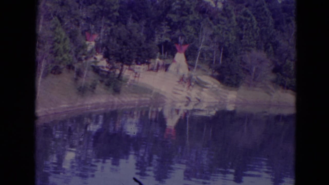1974 Orlando lakeside view with teepee-like structures among trees, sandy shoreline, and reflections across still water.