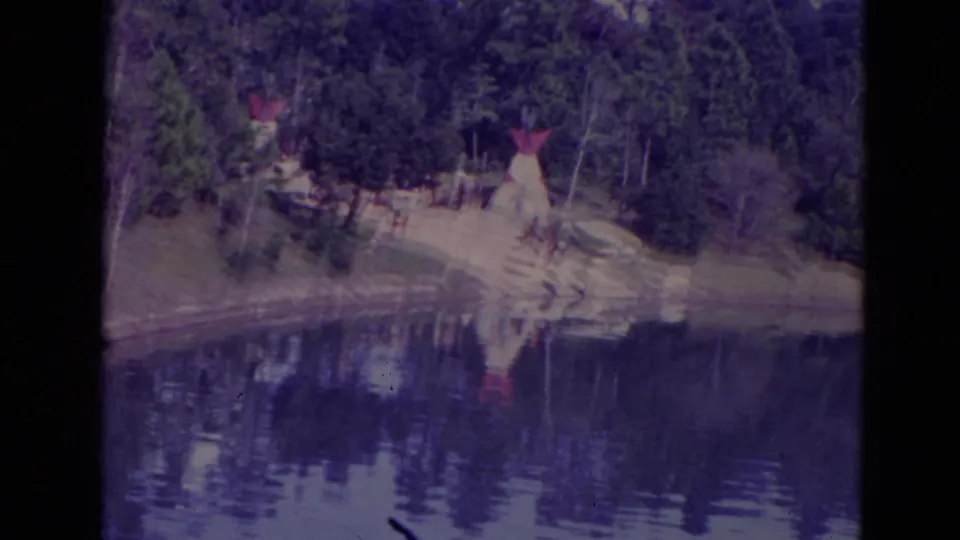 1974 Orlando lakeside view with teepee-like structures among trees, sandy shoreline, and reflections across still water.