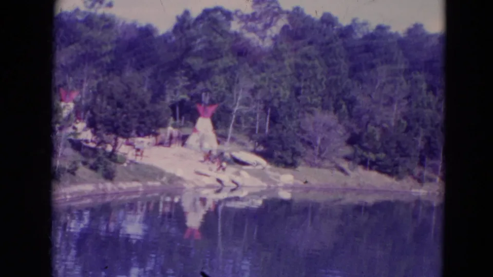 1974 Orlando home-movie frame with a wider view of teepee-like structures on a wooded shore beside dark reflective water.