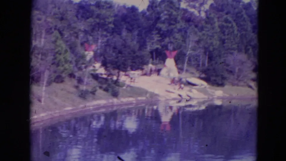 1974 Orlando frame showing teepee-like structures on a sandy wooded lakeshore, with reflections and blue-purple 8mm color shift.