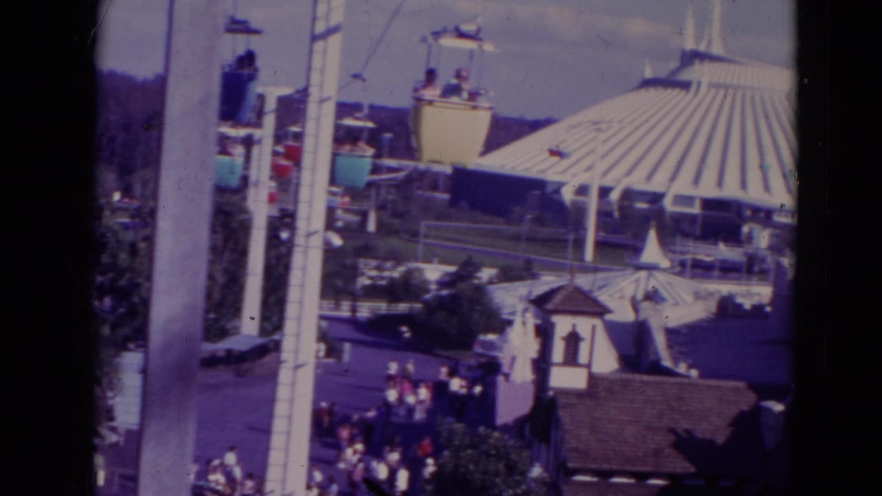 1974 Orlando home-movie frame showing an elevated view across white park buildings, a monorail beam, a ribbed pavilion, and small visitor crowds.