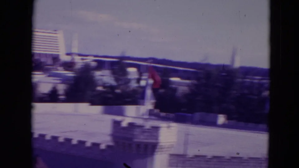 1974 Orlando home-movie frame with motion blur across a modern hotel, white park walls, treetops, rooftops, and distant spire shapes.