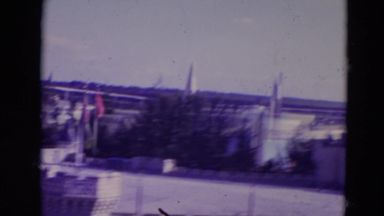 1974 Orlando home-movie frame showing castle-like spires beyond water, with flags, trees, pale sky, and visible 8mm frame borders.