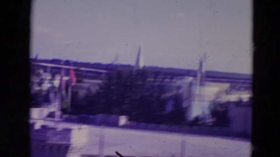1974 Orlando home-movie frame showing castle-like spires beyond water, with flags, trees, pale sky, and visible 8mm frame borders.