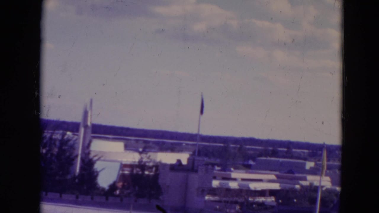 1974 Orlando 8mm frame with water, low park buildings, flagpoles, trees, and a distant horizon under a pale sky.