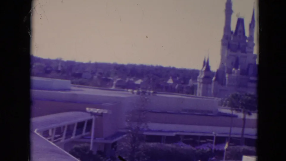 1974 Orlando home-movie frame showing castle towers behind low visitor buildings, railings, trees, and purple-shifted 8mm color.