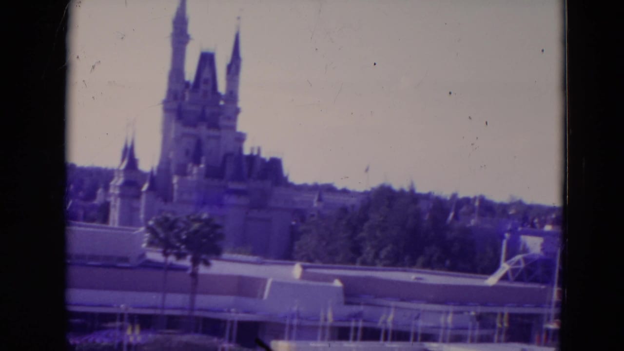 1974 Orlando home-movie frame showing Cinderella Castle in the distance with palm landscaping and park roofs in front.
