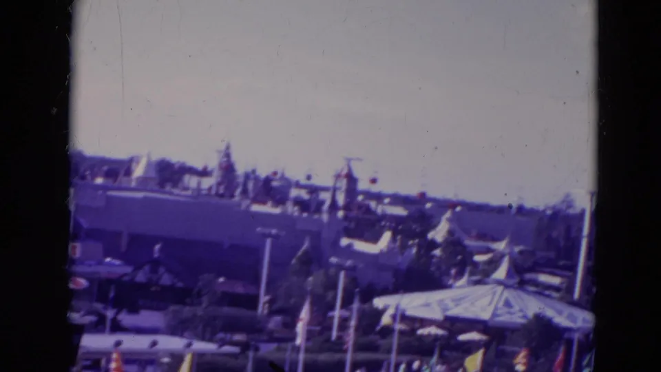 1974 Orlando home-movie frame showing park rooftops, flags, attraction structures, and landscaping during a pan.