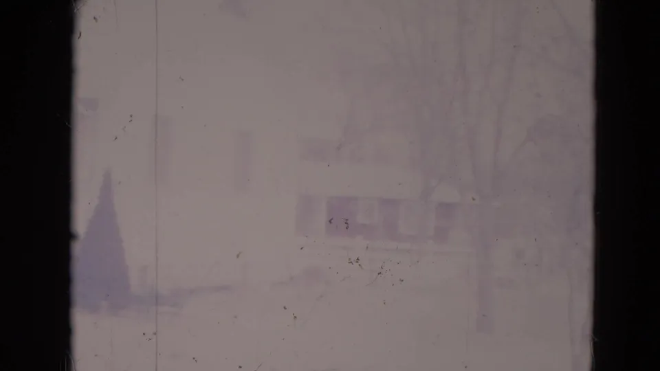1974 Orlando home-movie frame showing a pale suburban house exterior, bare trees, low shrubs, and a small evergreen through faded 8mm film.