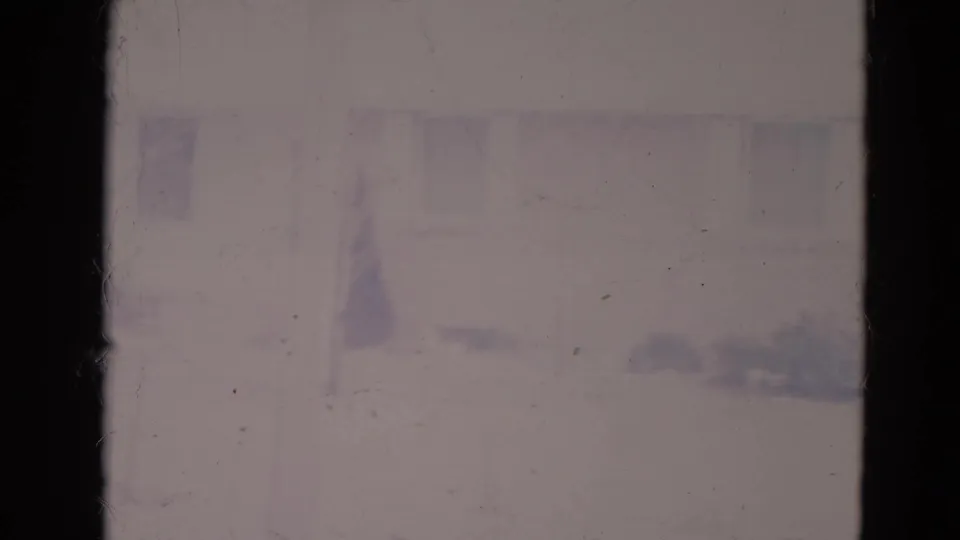 1974 Orlando home-movie still of a suburban house wall, faint windows, low landscaping, and a small evergreen softened by faded 8mm exposure.
