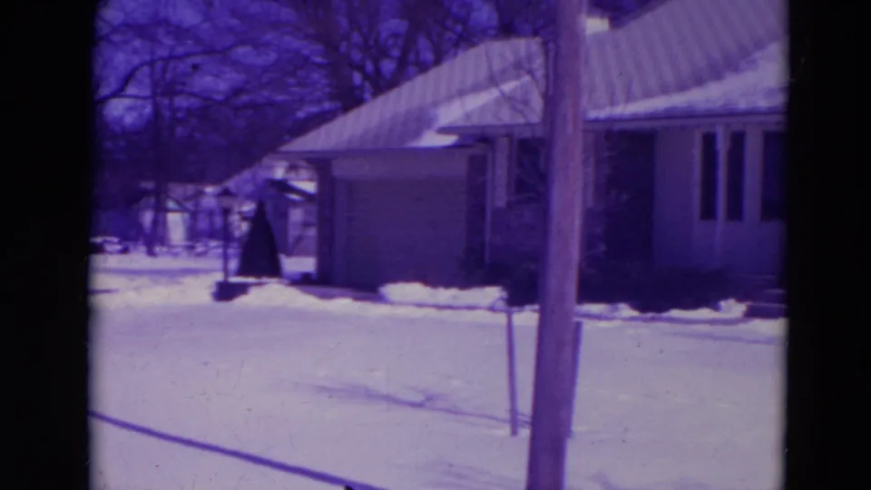 1974 Orlando frame showing the side of a snow-covered suburban house, a driveway or side yard, bare trees, and a foreground utility pole.