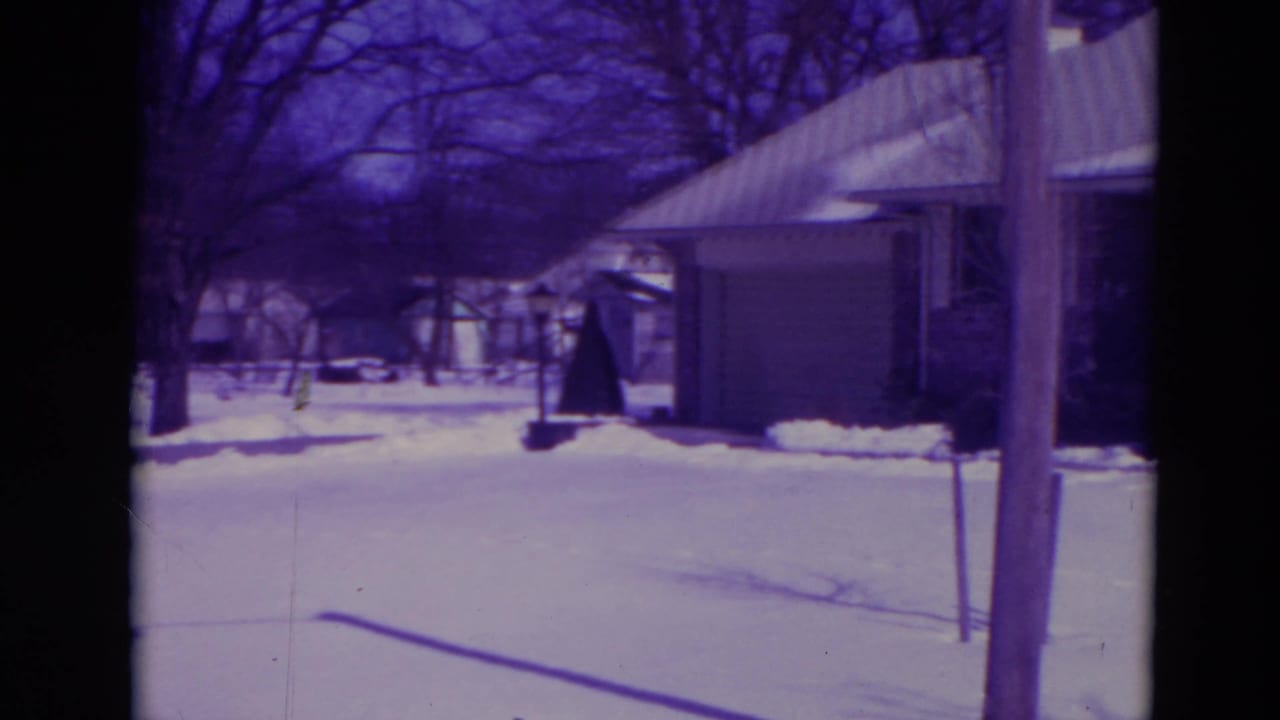Snow-covered yard with bare trees, a house and a detached garage beyond dark film borders.