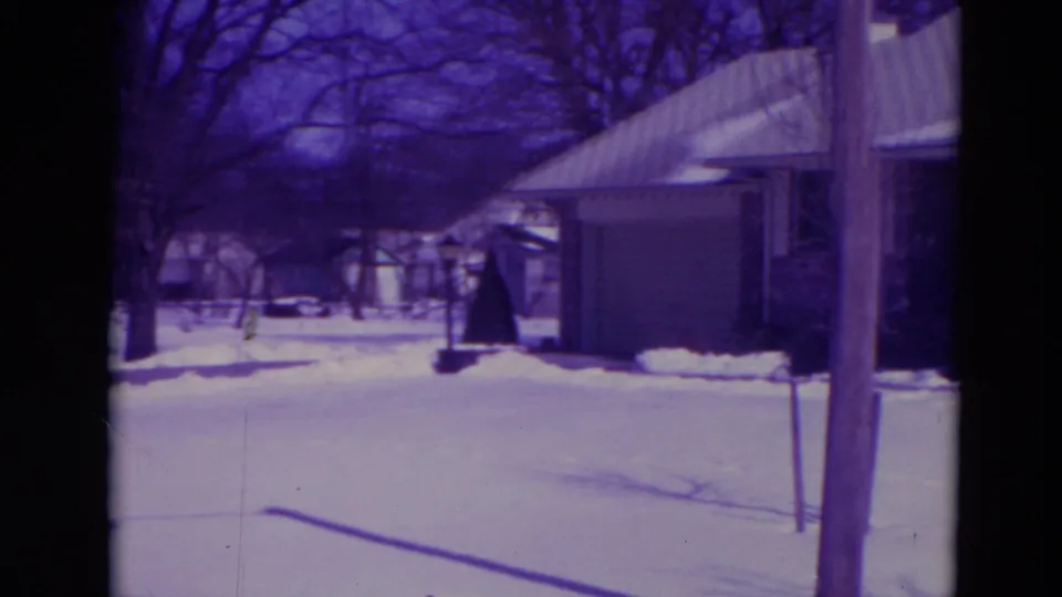 1974 Orlando home-movie frame showing a snowy residential yard, bare trees, a garage and a house in soft purple-tinted 8mm color.