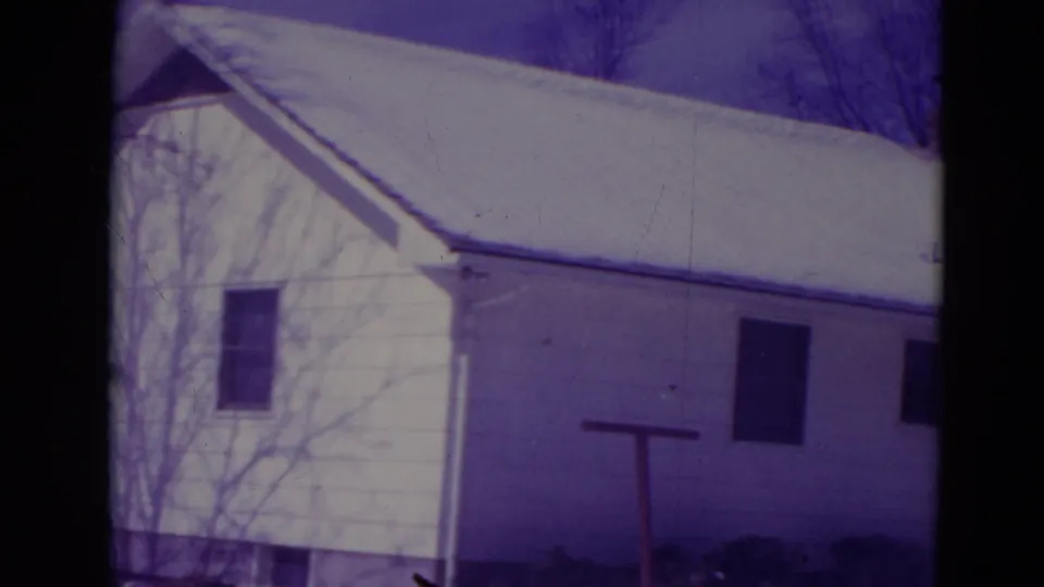 1974 Orlando home-movie frame showing a modest white house with snow on the roof and yard, bare trees, and aged 8mm color shift.